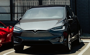 Tesla auto body shop Los Angeles: Dark gray Tesla Model X parked, red car partially visible, shop exterior in background.