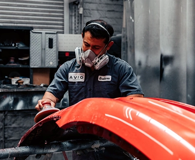 Auto Body Bumper Repair Los Angeles: Technician sanding a red car bumper, preparing for paint in a repair shop setting.